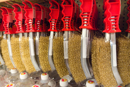 Metal brushes hang from their mounts, showcasing vibrant red handles and shiny bristles. Customers browse nearby, contemplating their options in the busy hardware store aisle.の写真素材