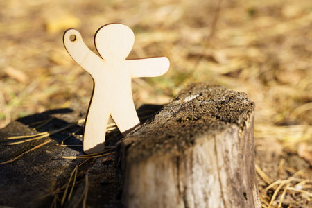 A wooden figure with arms outstretched is placed on a rustic tree stump amidst fallen leaves. Sunlight streams through the trees, casting playful shadows on the ground.の写真素材