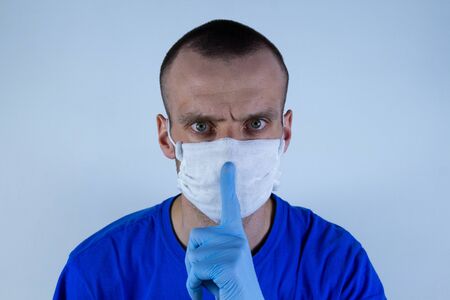 Young man in a protective mask on a blue background. Warns, Holds index finger in glove at mouthの写真素材