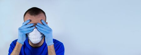 The doctor covers his face with his hands in gloves in a protective mask and blue clothes on a blue background. He is afraid of infection and does not know what to do and how to be.copyspace for textの写真素材