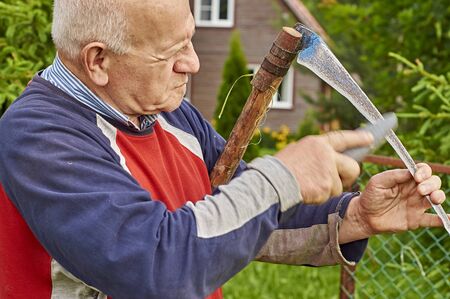 Senior man sharpening a scythe before mowing the grass in front of his summer residence                                の写真素材