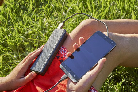 Young girl charging her smart phone with power bank sitting on green grass outdoorsの写真素材