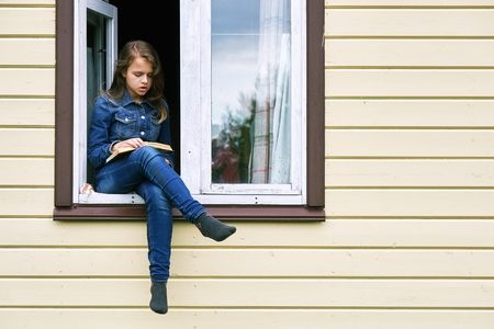 Girl reading a book on vacation sitting in the window of a country house with her legs dangling outsideの写真素材
