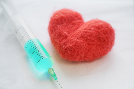 Red wool felted heart and a syringe with a blue medicine lying on a white bed sheet. Heart treatment conceptの写真素材