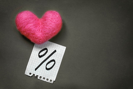 Pink wool toy heart lying with a notepad sheet with percent sign written on it on a dark grey backgroundの写真素材