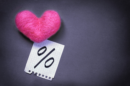 Pink wool toy heart lying with a notepad sheet with percent sign written on it on a dark violet backgroundの写真素材