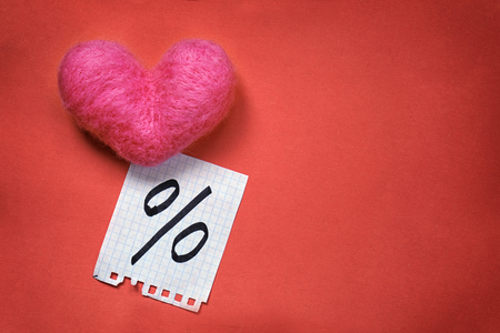 Pink wool toy heart lying with a notepad sheet with percent sign written on it on a dark orange backgroundの写真素材