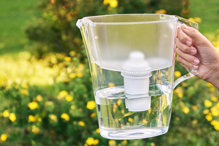 Girls hand holds a water filter jug in the sunny summer garden with yellow flowers on the backgroundの写真素材