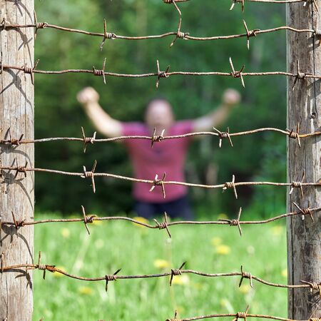 Fence with barbed wire on the foreground and an unrecognizable man with raised arms sitting on the...