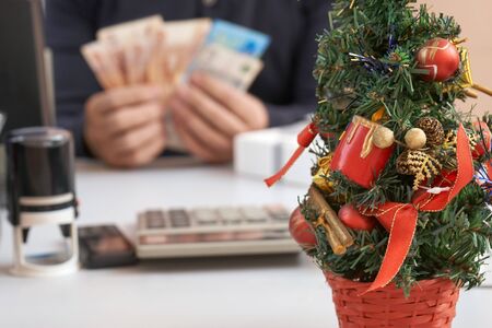Decotated small Christmas tree standing on the table on the foreground and a manager getsting a bonus money on workplace in Christmas or New Year eve on the backgroundの写真素材