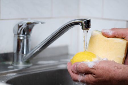 Male hands washing a lemon with a sponge in soap suds in a kitchen sinkの写真素材