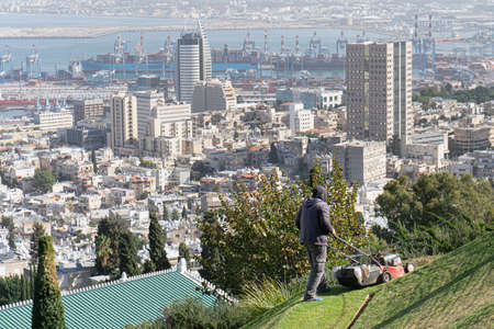 Haifa, Israel - November 28, 2019. Man mowing a lawn in Bahai gardens using a lawnmower on the Haifa downtown panorama background in sunny dayのeditorial素材