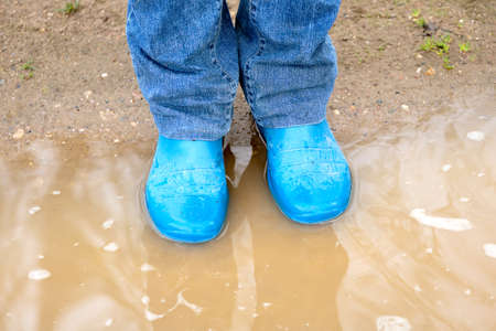 Feet of a woman wearing jeans in rubber boots standing in the dirty puddle on the rurual road