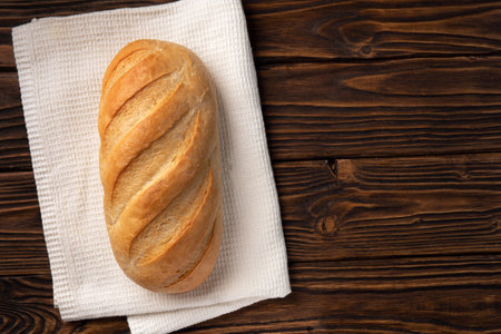 Loaf of a white bread on the white towel on the dark wooden boards