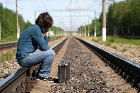 Woman with old suitcase sits on a rail of railroad and waits for a late train in summer morningの写真素材