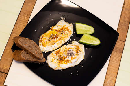 Fried eggs with cucumbers and rye bread in a black square plate. Top view.の写真素材