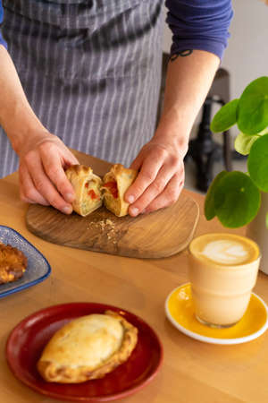 A bright image of baker's hands breaking open a freshly baked cheese and pepper empanada. Situated...