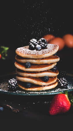 Dusted icing sugar on a stacked tower of pancakes topped with summer fruits including strawberries, blueberries and blackberries. Styled on a plate on a dark backgroundの写真素材
