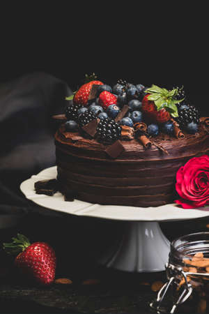 Delicious Double Chocolate Cake Dessert topped with Chocolate Flakes and Summer Fruits, including blackberries, strawberries and blueberries. Styled on a white cake stand with a rose and jar of almondの写真素材
