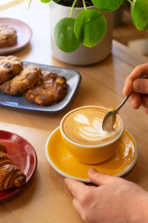 A spoon in a yellow cup and saucer of cappuccino coffee with freshly baked savory and sweet treats...