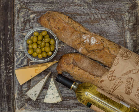 a bottle of white wine, olives, two pieces of cheese and a pair of rye baguettes in a paper bag on a...