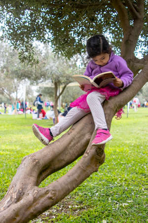 Cute little girl reading a book on a tree in a city parkの写真素材
