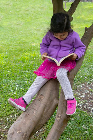 Cute little girl reading a book on a tree in a city parkの写真素材
