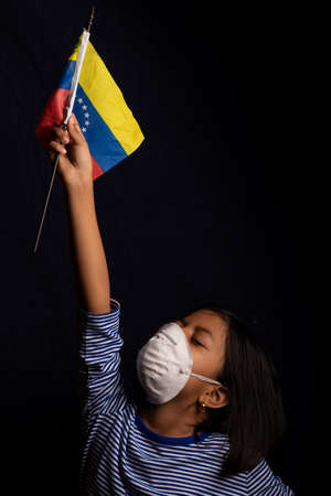 Portrait of little Venezuelan girl wearing medical mask and holding hopefully the flag of Venezuelaの写真素材