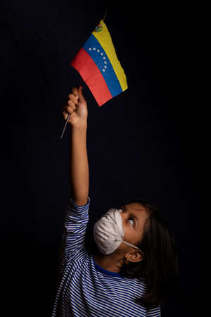 Portrait of little Venezuelan girl wearing medical mask and holding hopefully the flag of Venezuelaの写真素材