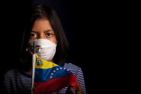 Portrait of little Venezuelan girl wearing medical mask and holding hopefully the flag of Venezuelaの写真素材