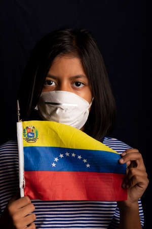 Portrait of little Venezuelan girl wearing medical mask and holding hopefully the flag of Venezuelaの写真素材