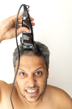Handsome gray-haired man is cutting hair himself with an electric hair clipper.