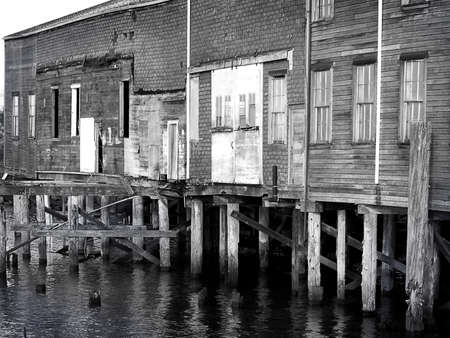 Old Mill buildings stand vacant on river pilings as water laps beneath in this lonely black & white image.の写真素材