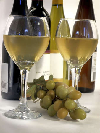 Two tall glasses of white wine with bunch of white grapes and bottles in background.の写真素材