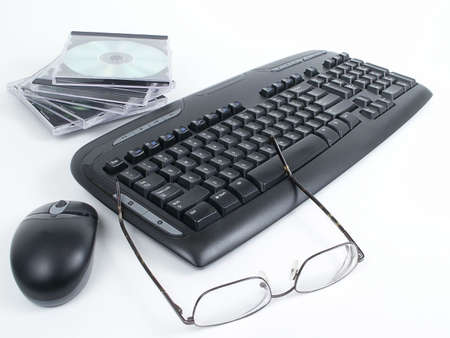 Black wireless keyboard and mouse with stack of CDs in cases, and man's glasses in foreground, isolated on white background.の写真素材