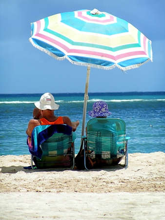 Older couple sit under umbrella on warm tropical beach.の写真素材