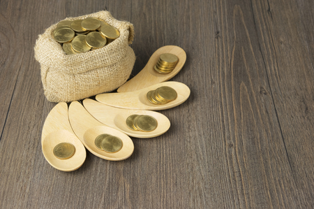 Gold coins in wooden spoon with brown sack of gold coins on wood background.の写真素材