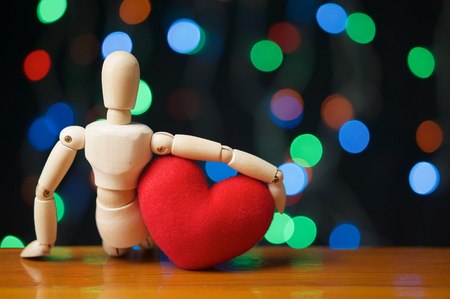Wooden dummy hug red heart shape and sit on wooden table have colorful bokeh as background.の写真素材