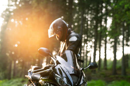 Young male Biker with racing motorbike on forest country road having a break during the sunset.の写真素材
