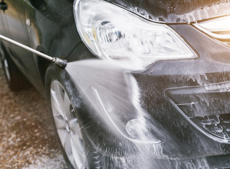 Front of black car gets cleaned by steam radiator at DIY car wash. Nice sunlight, water mist and foam.の写真素材