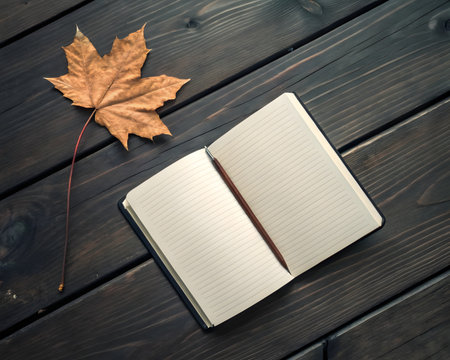 An open notebook rests alongside a maple leaf on a dark wooden surface, capturing the essence of the autumn season with its warm colors and natural textures.の素材