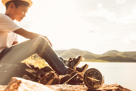 young asian boy sitting alone with compass at mountain road at sunset skyの写真素材