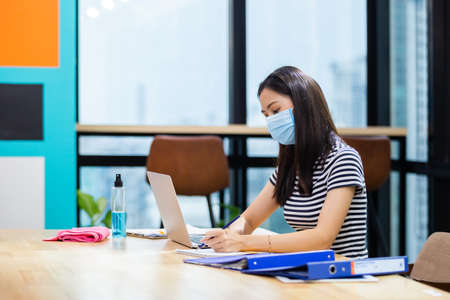 Asian women wear masks protect against airborne disease and salivary infections, during outbreak of Covid 19 virus (Coronavirus) and working with laptop in office.の写真素材