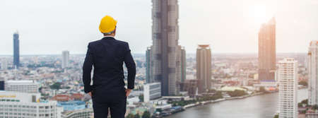 architect man wear black suit and yellow helmet view from back standing on building looking for...
