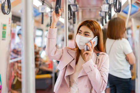 Asian women passenger wearing medical face mask for prevent dust and protect coronavirus pandemic, holding smart mobile phone standing in subway train transit system public.の写真素材