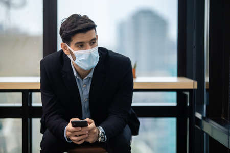 handsome businessman wear masks protect against airborne disease and salivary infections, during outbreak of Covid-19, and holding smartphone sitting in office.の写真素材