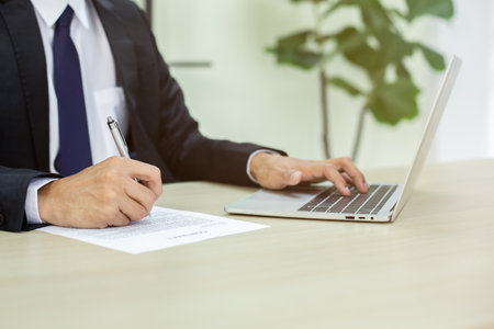 Close up hand of businessman holding pen and writting on paperwork using laptop in office. business...