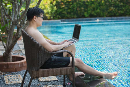 asian businessman seat a chair working with laptop in swimming poolside. work new normal. lifestyle travel in holiday vacation. relaxation concept.の写真素材