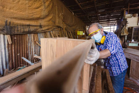 asian senior man carpenter wear protective mask dust and headphone working to measure size of wood....