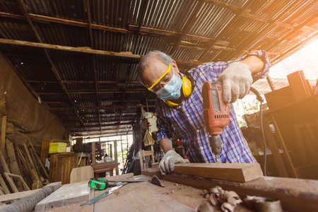 asian senior man carpenter wear protective mask dust and headphone working with electrical drill to...
